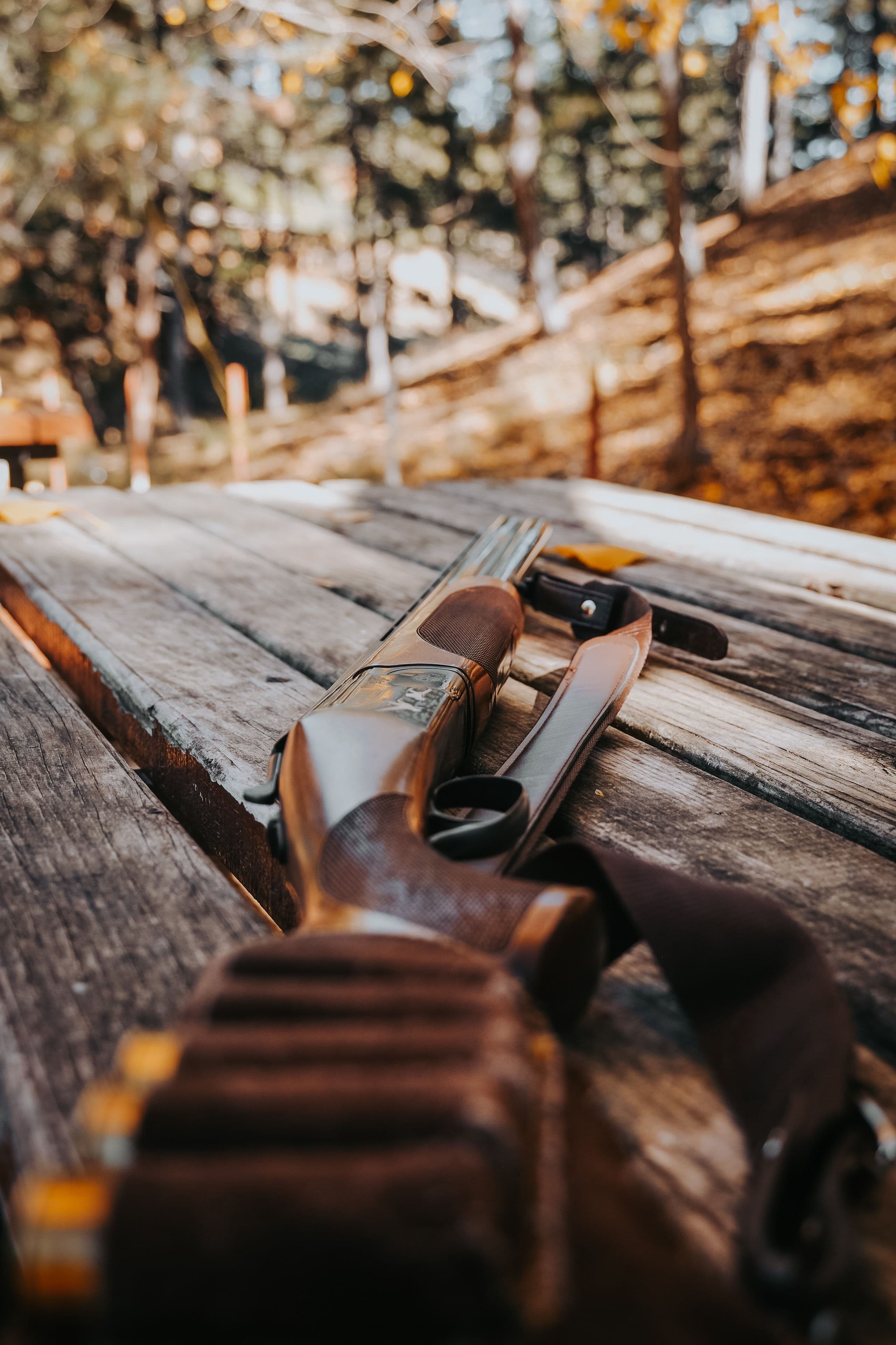 A shotgun with a leather sling rests on a rustic wooden table in the forest.